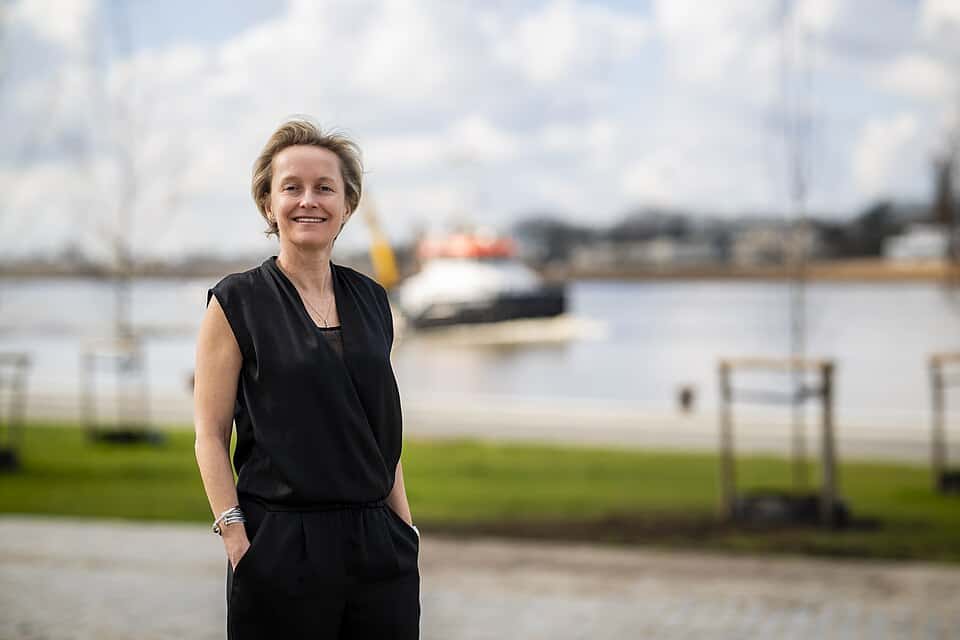 Minister De Ridder bij Woumen met water en boten op achtergrond.
