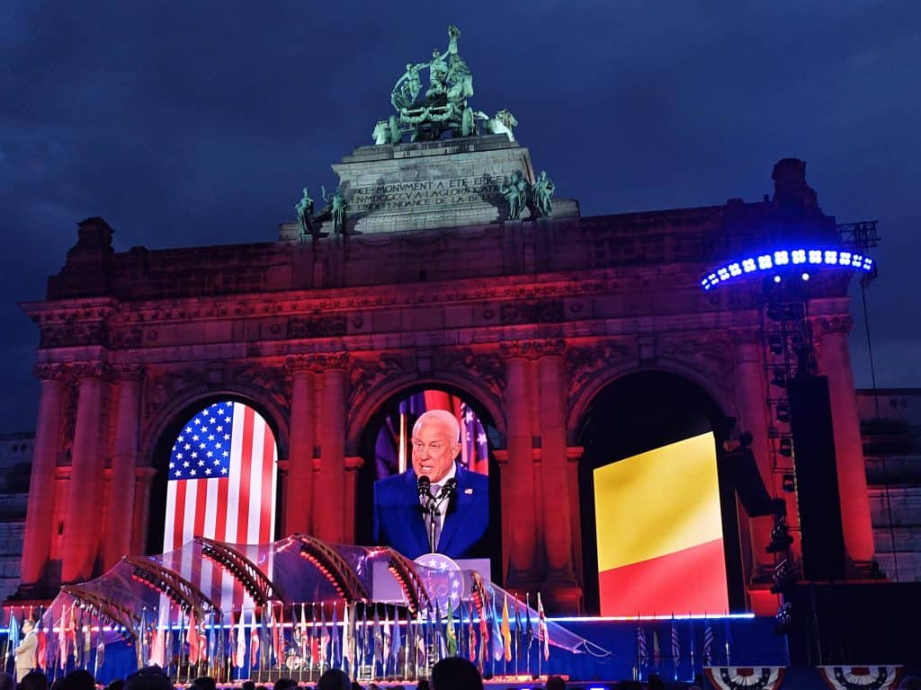 Amerikaanse vlag en een man spreekt op een groot scherm tijdens viering in Brussel.