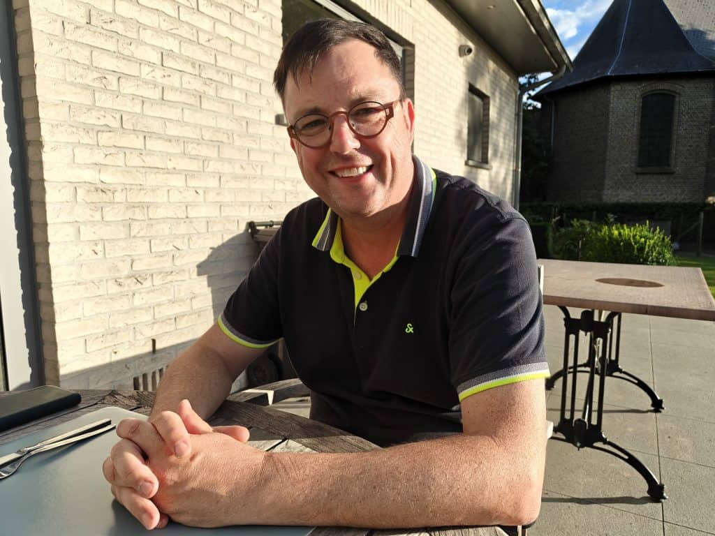 Man met bril en glimlach zit buiten aan tafel.